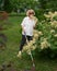 An elderly blind woman smells a flowering shrub while walking in the park.