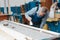 An elderly cabinetmaker in overalls and glasses paints a wooden board with a brush on a workbench in a carpentry shop.