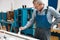 An elderly cabinetmaker in overalls and glasses paints a wooden board with a brush on a workbench in a carpentry shop.