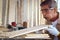 Elderly carpenter wearing safety glasses goggles blowing sawdust off piece of wood plank, senior craftsman working at wooden desk