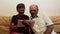An elderly couple is sitting on a sofa at home and watching photos on a tablet pc, smiling and talking actively.