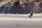 An elderly couple walk across an empty beach at the base of tall cliffs while holding hands