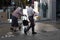 Elderly couple with wife bent over walker and her husband somewhat casually walk past a children\\\'s shop