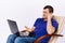 An elderly deaf man in a leather chair with a laptop and a smile shows a hearing aid at the camera. Online health