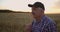 An elderly farmer man in a shirt and baseball cap stands in a field of cereal crops at sunset and looks at the spikes of