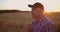 An elderly farmer man in a shirt and baseball cap stands in a field of cereal crops at sunset and looks at the spikes of