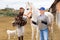 Elderly female stable keeper leading horse to outdoor riding arena while Asian woman holding saddle