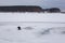 Elderly fisherman in dark clothes fishing on winter fishing rod on frozen river