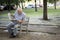An elderly gray-haired man carefully reads a newspaper while sitting on a bench in the park