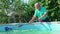 An elderly gray-haired man cleans the walls of the pool with a mop.