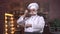 An elderly male chef straightening his thick gray mustache while standing in the kitchen