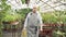 An elderly man in an apron with tools and a watering can walking in the greenhouse. Agriculture, farming and gardening