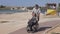 An elderly man carries a woman in a wheelchair along the sea