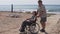 An elderly man carries a woman in a wheelchair along the sea