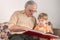 An elderly man in glasses reads a book to his grandson. The child, listens carefully