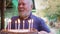 An elderly man with a gray beard blows out the candles on the birthday cake.