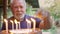 An elderly man with a gray beard blows out the candles on the birthday cake.