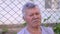 Elderly man with a gray mustache and hair dozing, leaning against a metal mesh fence. Outdoors, summer day