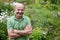 An elderly man with a mustache and a bald spot in a green T-shirt is standing among flowers in the summer garden, arms