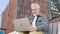 Elderly man opening laptop while sitting on outdoor bench