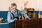 An elderly man paints wooden boards with dark varnish in a carpentry workshop. A pensioner in a medical mask