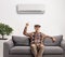 Elderly man seated on a sofa pointing up towards an air conditioning unit