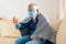 An elderly man sick with a fever and using a thermometer at home, sitting on the couch with a virus mask