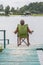 An elderly man sits in a tourist chair on a wooden platform with a lake in the background.  Concept: tourism and travel, retiremen