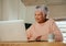 Elderly multi-ethnic female sitting at home counter using laptop, smiling.