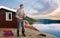 An elderly Norwegian is standing on a rock by the water and fishing.