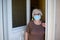 Elderly old senior woman opening front door of house and welcoming people at home wearing a surgical mask to avoid covid19 and cor