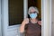 Elderly old senior woman opening front door of house and welcoming people at home wearing a surgical mask to avoid covid19 and cor