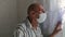 An elderly retired man in a medical mask stands near the window of a house and looks out. Elderly people in quarantine amid the co