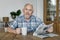 Elderly retired man with newspaper seated at table with cup of tea in kitchen