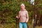 Elderly sixty-year-old woman in sportswear is engaged in Nordic walking with sticks in the forest in the autumn