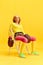 Elderly sportive woman in colorful uniform sitting on chair in retro boxing gloves, posing against yellow studio
