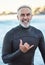 Elderly, surfer and beach with hand sign for portrait with smile in summer by ocean for wellness, fitness and health