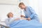 Elderly woman lying in the hospital room bed, nurse holding the needle to connect the IV to the patient`s arm