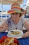 Elderly woman in  multicolored hat over plate of noodle soup at waterfront cafe