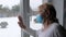 Elderly Woman In Protective Face Mask Standing At Window Waving Her Hand