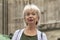 An elderly woman with White gray Hair stands against the background of an ancient building.