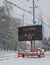 Electric road traffic mobile sign by the side of a snow covered road with snow falling warning of black ice on road.