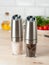 Electric spice mills made of metal. On a light wooden background. In the background is a plate with vegetables and olive oil