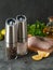 Electric spice mills made of metal. Spices and lemon dolly are scattered nearby. In the background chicken fillet, herbs and olive