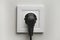 Electric white socket and one plugged in power cord on white wall background