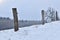 Electric wire fence, wooden pile with isolator and snow. Agricultural landscape, snowy farmland in winter