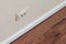 Electrical outlet on beige home wall with wallpaper, white baseboard and brown wooden laminate