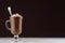 Elegance hot chocolate in classic irish coffee glass with marshmallows and silver spoon on dark brown wall and white wooden table.