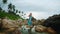 Elegant lady in blue gown stands on rock amidst serene tide pool, flanked by boulders under cloudy skies. Reflective