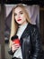 Elegant positive blond woman with trendy makeup bright vibrant red lips in leather coat drinking coffee from red disposable cup